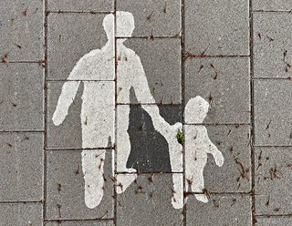 Image shows photo taken from above, of a pavement. The pavement has an image painted on it where a tall person and a small person walk together.